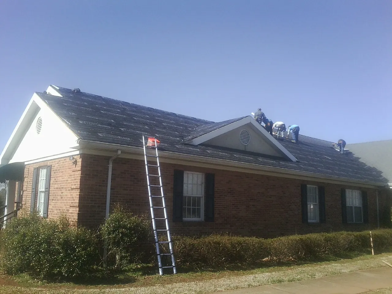 Crew tearing shingles off a brick ranch roof with a ladder set — Emergency Roof Repair work by our crew in Bethune